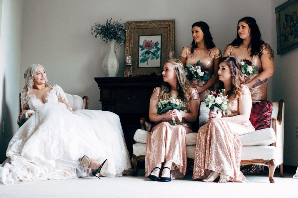 A bride in a lace dress and bridesmaids in sequin gowns share a candid moment indoors.